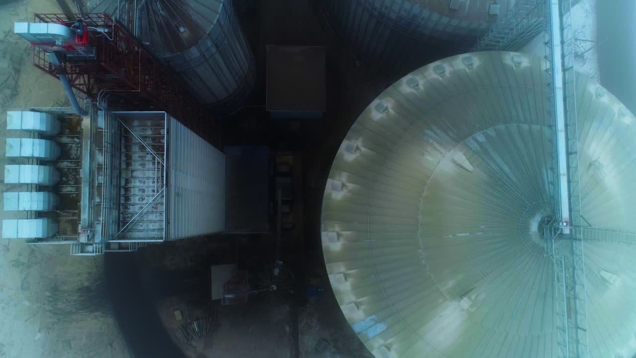 Flying above the diverse in size elevator constructions and premises. Huge tanks of modern agribusiness plant on foggy grey day. Top view.