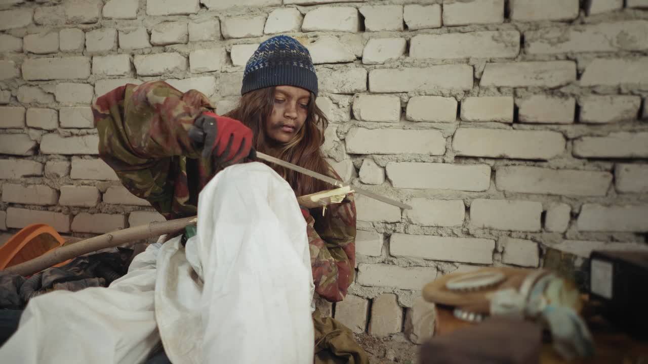 Un niño náufrago con pantalones blancos sucios y chaqueta de camuflaje afila un trozo de madera grueso con un machete grande mientras está sentado cerca de una pared de ladrillo, junto a utensilios de cocina esparcidos y una olla en un hornillo dentro de un refugio frío y descuidado.