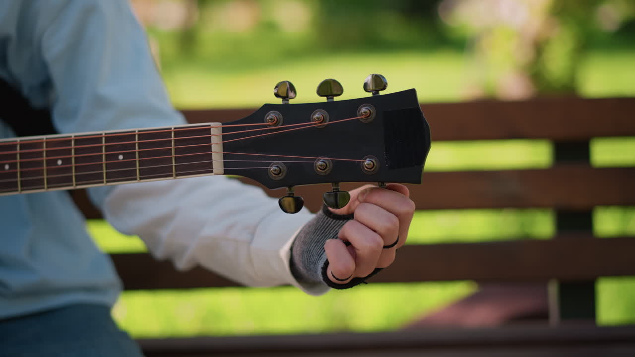 Primer plano de la mano de un guitarrista blanco afinando el clavijero de la guitarra, ajustes precisos de las clavijas, colocación y calibración de los dedos para el tono, ambiente tranquilo de tutorial al aire libre con destreza y técnica enfocadas.