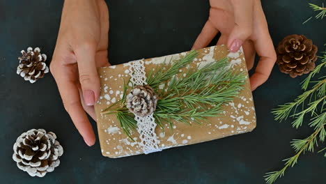 female hands shows a wrapped gift decorated with a pine cone, a branch and painted snow on a table