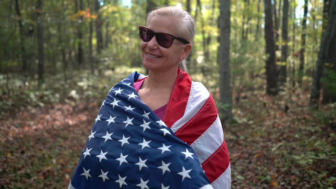 el primer plano de una mujer rubia y bonita envolviendo una bandera americana a su alrededor y sonriendo en un bosque iluminado por el sol