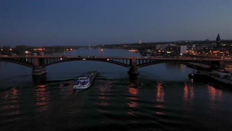 Drohnenaufnahme-Rund-Um-Die-Altstadt-Von-Mainz,-Die-Die-Stadt-Im-Hintergrund-Zur-Magischen-Stunde-Am-Abend-Mit-Einem-Schiff-Auf-Dem-Rhein-Zeigt