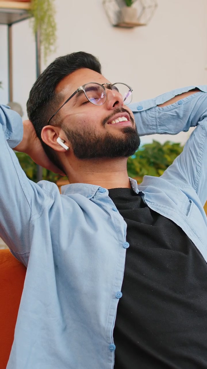 Uomo indiano felice e rilassato che si riposa sul divano di casa con auricolari wireless, ascoltando musica ambient.