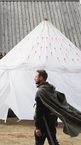 skillful medieval warrior walks about territory of settlement on cloudy day. wind blows around long cloak of young bearded knight side view