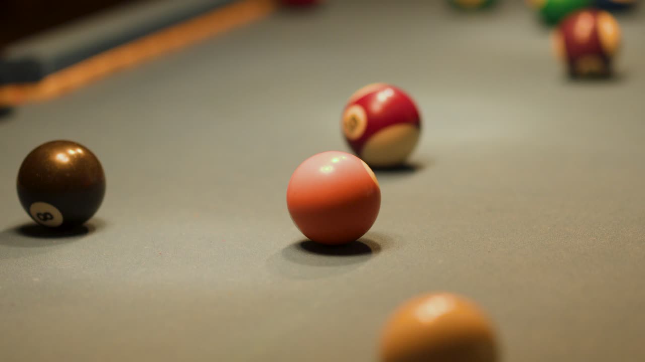Multiple billiard balls collide and roll on a felt table under warm, low indoor lighting