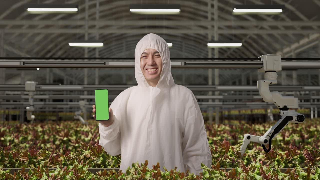 азиатский исследователь улыбается и показывает зеленый экран смартфона камере, стоя в теплице с умными роботами-фермерами
