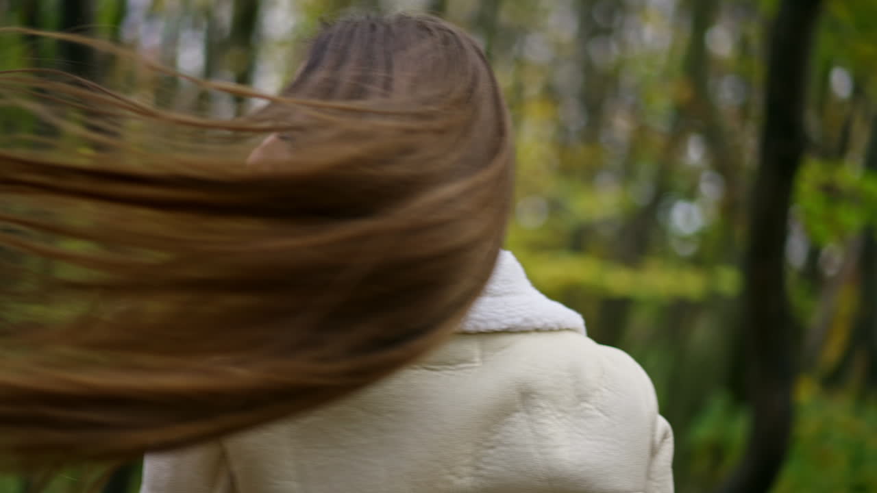 Beautiful lady waves her long dark hair in front of camera. Woman in white jacket walking by the autumn colorful park.