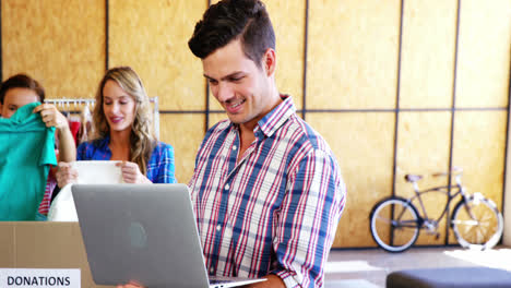 Hombre-Trabajando-En-Una-Computadora-Portátil-Mientras-Los-Voluntarios-Clasifican-La-Ropa-En-El-Fondo