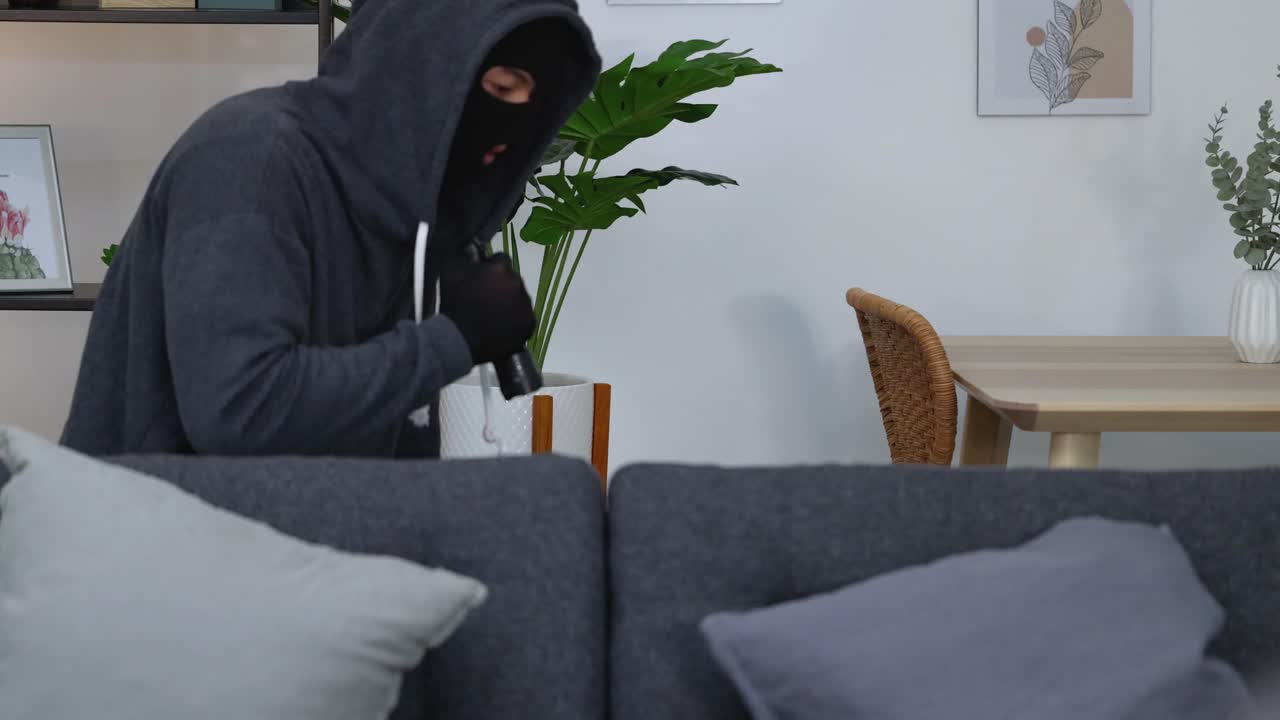 A criminal in a balaclava uses a flashlight to search a dark living room