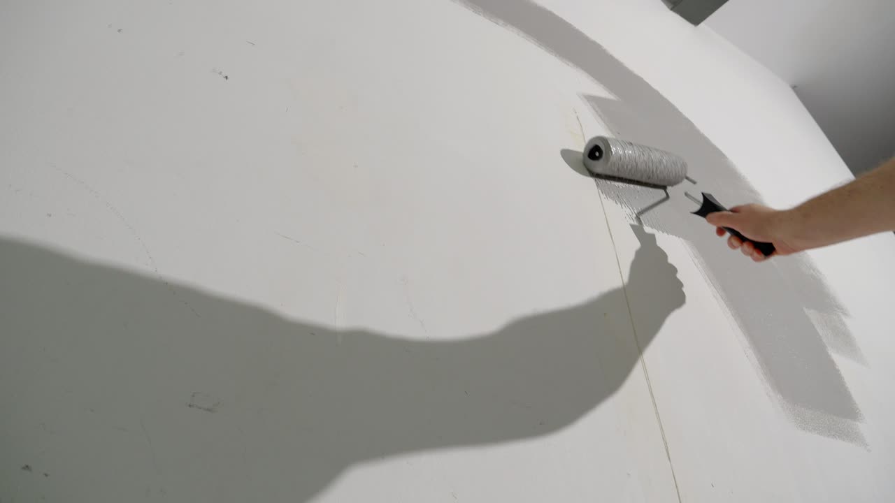 un hombre está pintando una pared blanca de gris con un rodillo de pintura, disparado desde abajo