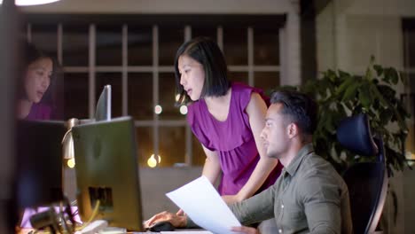 Focused-diverse-male-and-female-colleague-at-desk,-talking-and-using-computer-at-night-in-office