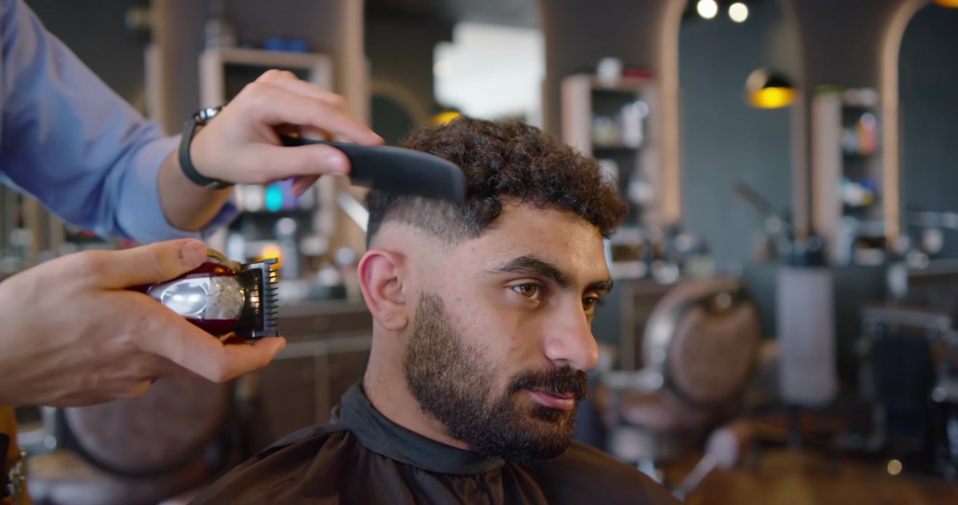 Barbiere che taglia i capelli a un uomo afroamericano seduto, creando una sfumatura a pelle usando il tagliacapelli in un salone da barbiere.