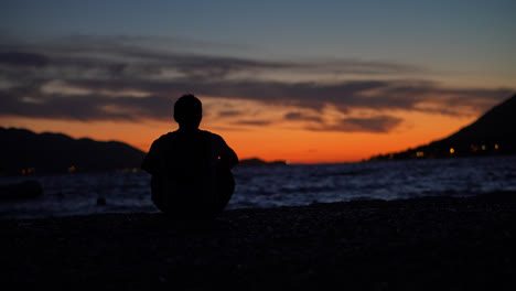 Young-male-sitting-alone-on-a-sandy-beach-watching-a-beautiful-orange-sunset-over-the-tropical-ocean-in-Croatia,-Europe