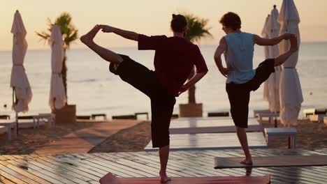 Dos-Chicos-Están-Parados-Sobre-Una-Pierna-En-La-Playa-Y-Meditando.-Clase-De-Yoga-Estilo-Zen-En-La-Playa-Por-La-Mañana.