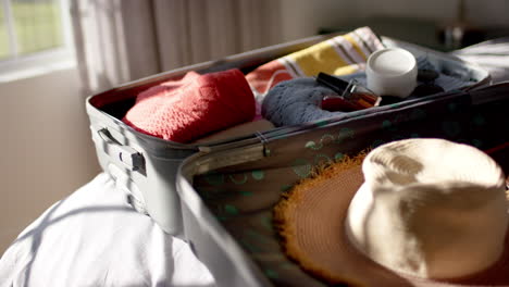 a suitcase lies open on a bed, filled with clothes and a straw hat