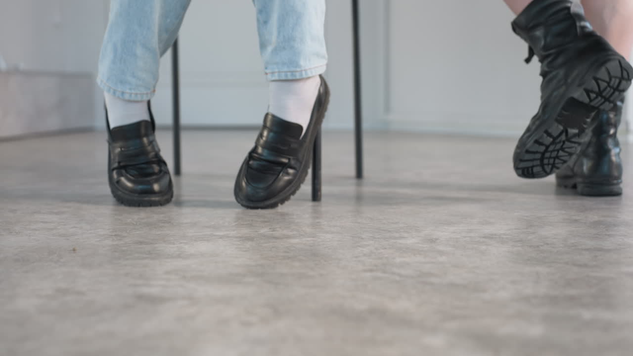 low angle close up view of fair skinned person wearing black combat boots walking behind seated individual in blue jeans and black loafers against softly lit modern interior background