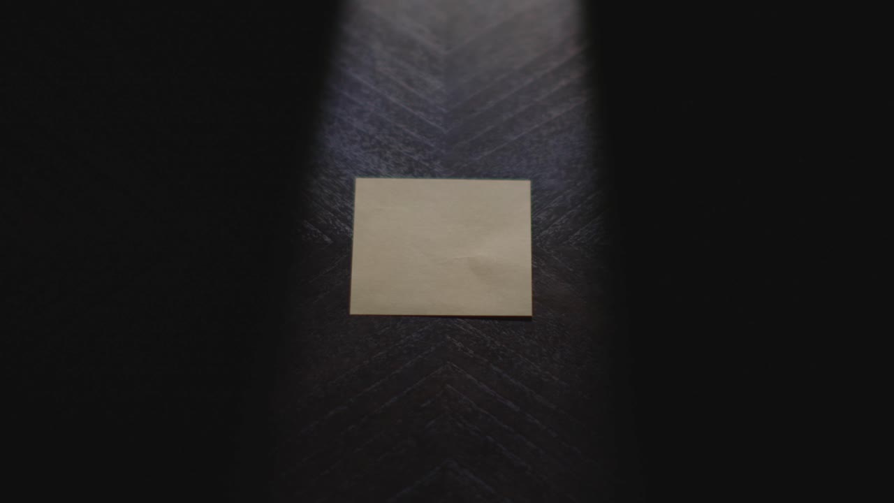 A close up of "Saturday" being written on a Post it Note. The note is then picked up and placed back down. Shot in dramatic lighting and slow motion