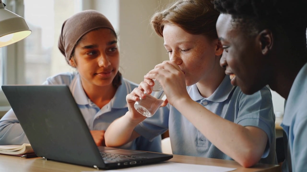 Studerende i midten rækker ud og løfter vandflaske, holder pause for at drikke i skolen med bærbar computer, iført blå poloer.