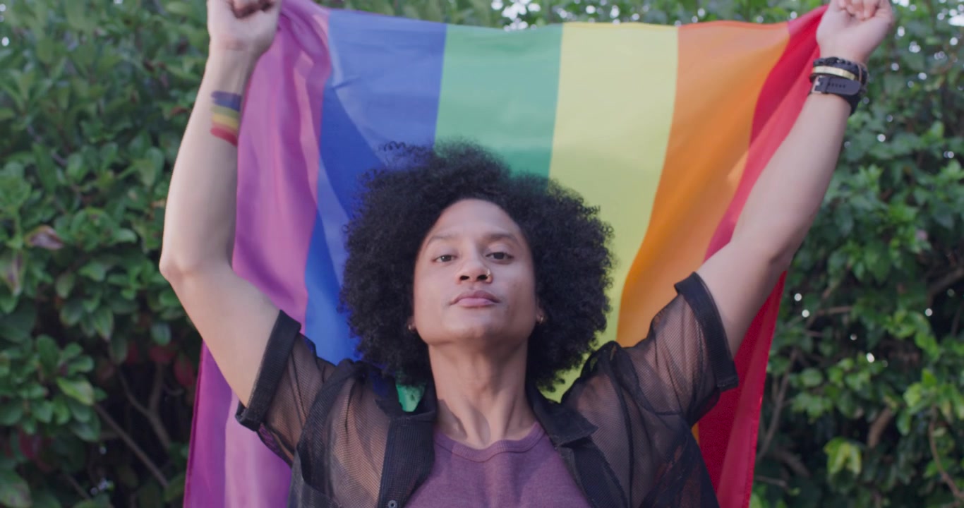 Homem afro-americano abrindo a camisa transparente, estendendo a bandeira do arco-íris acima da cabeça, mostrando orgulho no quintal.