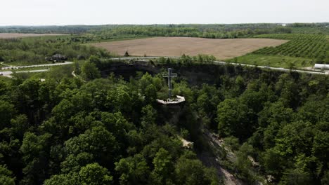 aerial orbit shot of cross standing on hill surrounded by green forest trees at devil's punch bowl, ontario