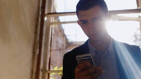 Unhappy-Young-Businesswoman-Writing-Sms-Sitting-In-An-Abandoned-House-Near-The-Window-The-Sun's-Rays