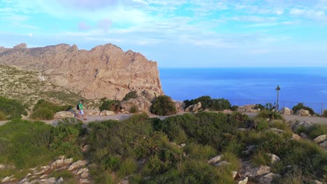 aerial tracking shot of a young man walking along a coastal path at mirador de es colomer, mallorca. dramatic cliffs and ocean views in scenic mediterranean light