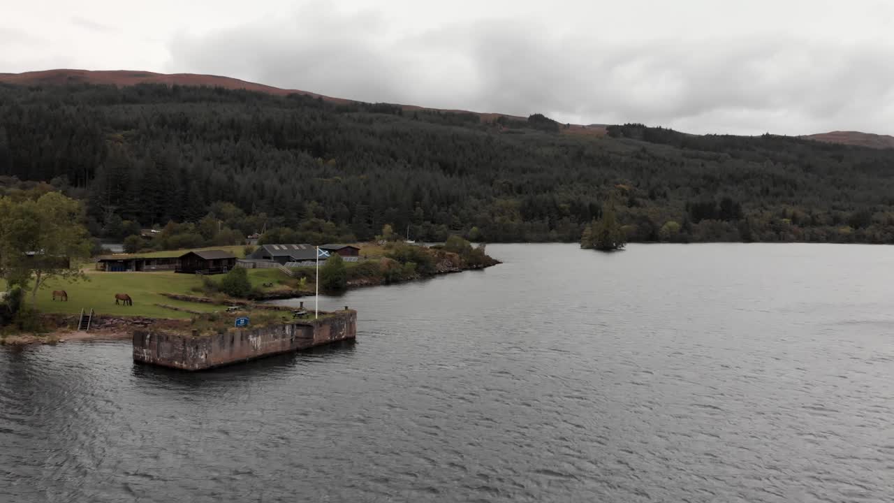 la bandera escocesa ondea desde el muelle en el lago ness mientras los caballos pastan cerca, aéreo