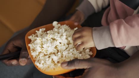 African-American-father-spending-time-with-small-son-sitting-on-couch-eating-popcorn-watching-TV,-close-up