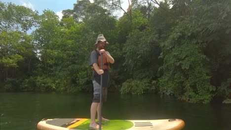 Ein-Bärtiger-Rothaariger-Mann-Draußen-Am-Ufer-Des-Nils-Im-Tropischen-Afrika-Auf-Einem-Stand-up-paddle-board