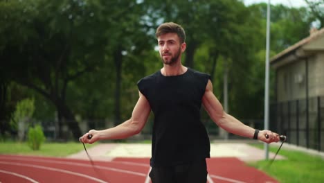 Hombre-Guapo-Y-Musculoso-Parado-En-La-Pista-De-Atletismo-Al-Aire-Libre-En-Un-Estadio-Moderno-Haciendo-Ejercicio-Con-Cuerda-Para-Saltar