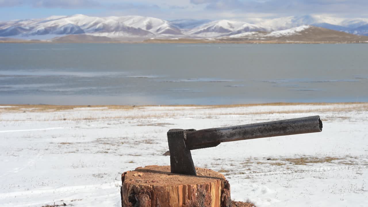 An axe is left in a tree stump, a vital tool for chopping firewood in the harsh Mongolian winter. In the background, a lake and snow-covered mountains complete the scene