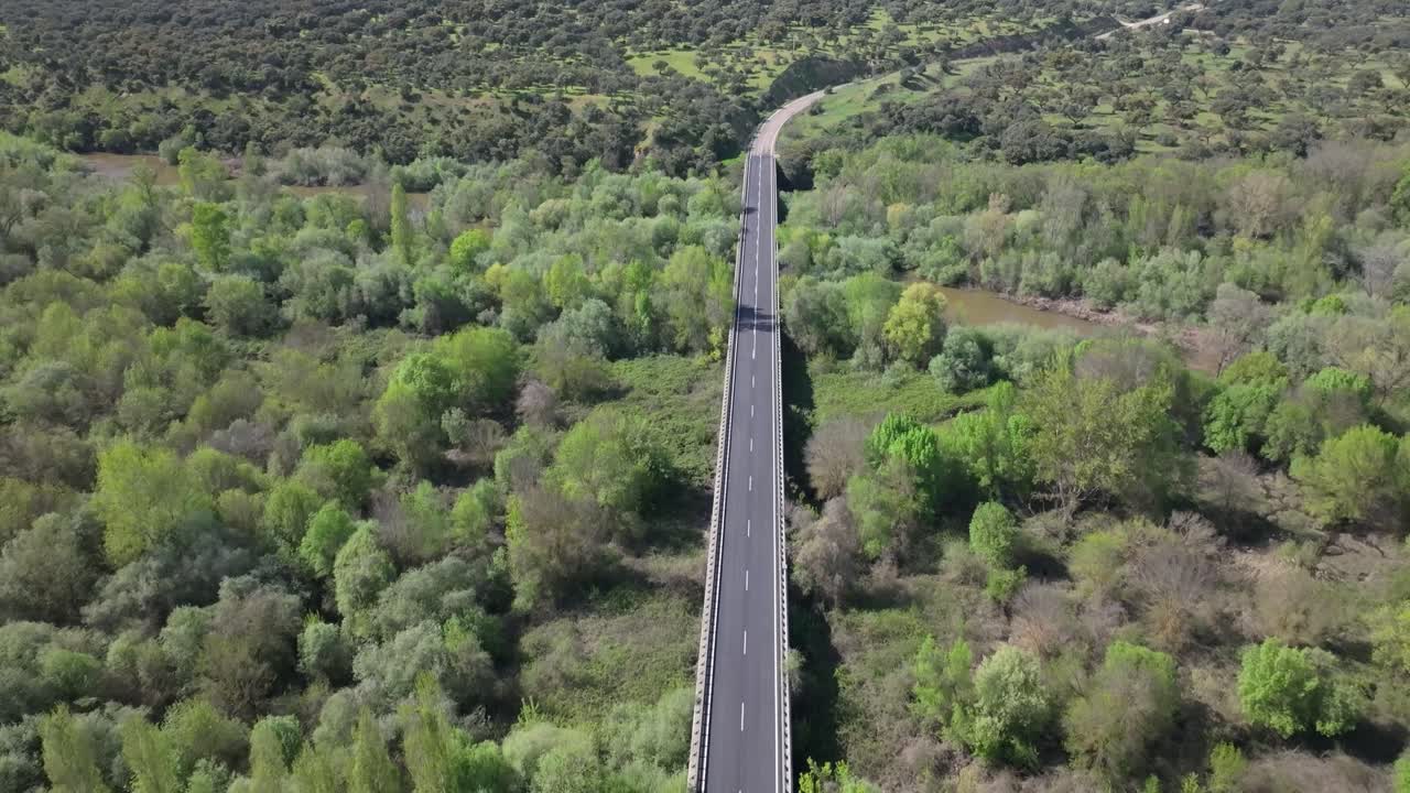 Reverse drone flight over a road where asphalt changes color due to a bridge spanning a wide muddy river that splits into two arms. Lush green surroundings and sediment-filled brown water visible.