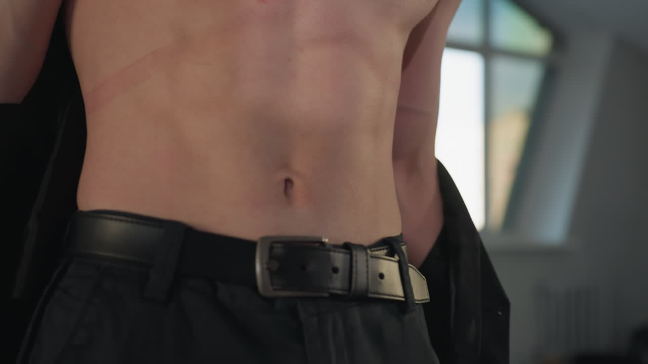 muscular man in bright room removes sleek black waistcoat slowly revealing toned torso while morning city light streams through large window, closeup highlights buttons strong arms