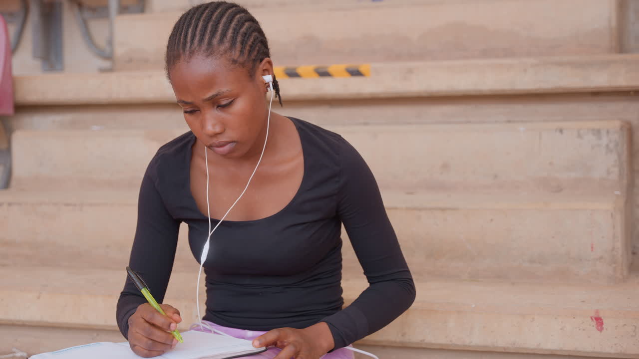 Studente che prende appunti. Studentessa di colore che annota appunti all'aperto. Donna con auricolari che studia sui gradini del campus. Donna afroamericana che scrive diligentemente su un quaderno durante una sessione di studio all'aperto.