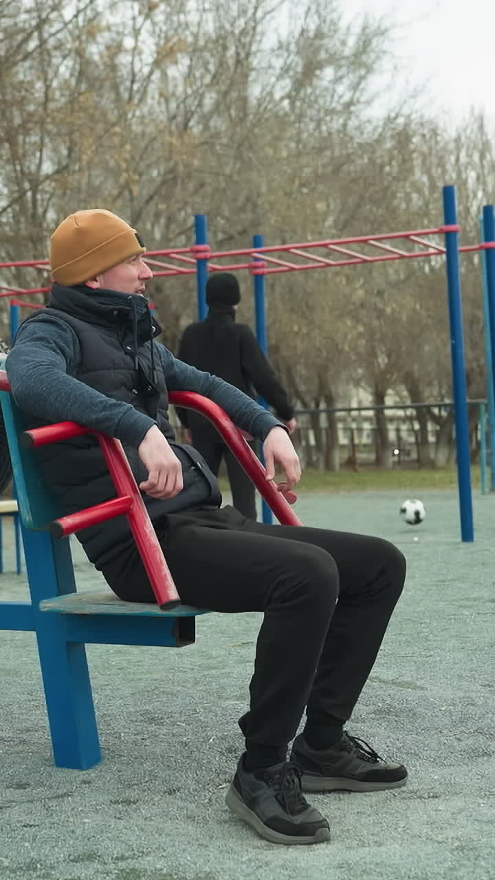 A coach leans back on workout equipment, casually shaking his leg, in the background, two boys are actively playing football