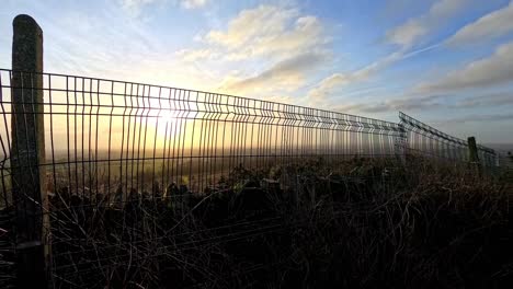 Golden-sunrise-clouds-time-lapse-passing-over-metal-fenced-farmland-meadow-in-morning-daylight