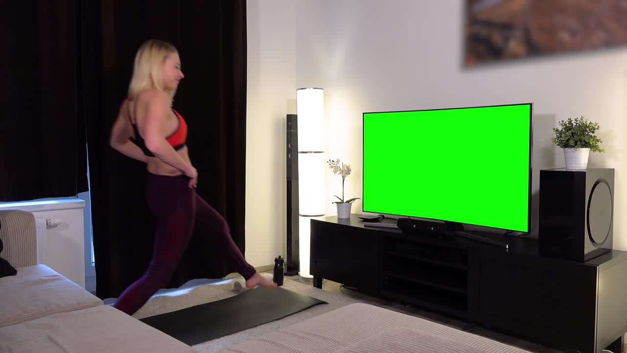 A young beautiful woman does lunges in front of a TV with a green screen in an apartment