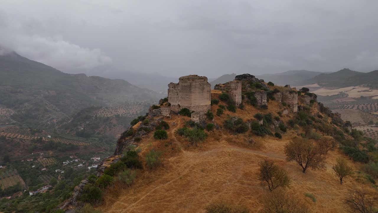 Aerial tracking back from a hilltop castle, Turón Castle ruins in Ardales, Málaga, Spain, revealing weathered stone walls and expansive farmland under a moody, overcast sky