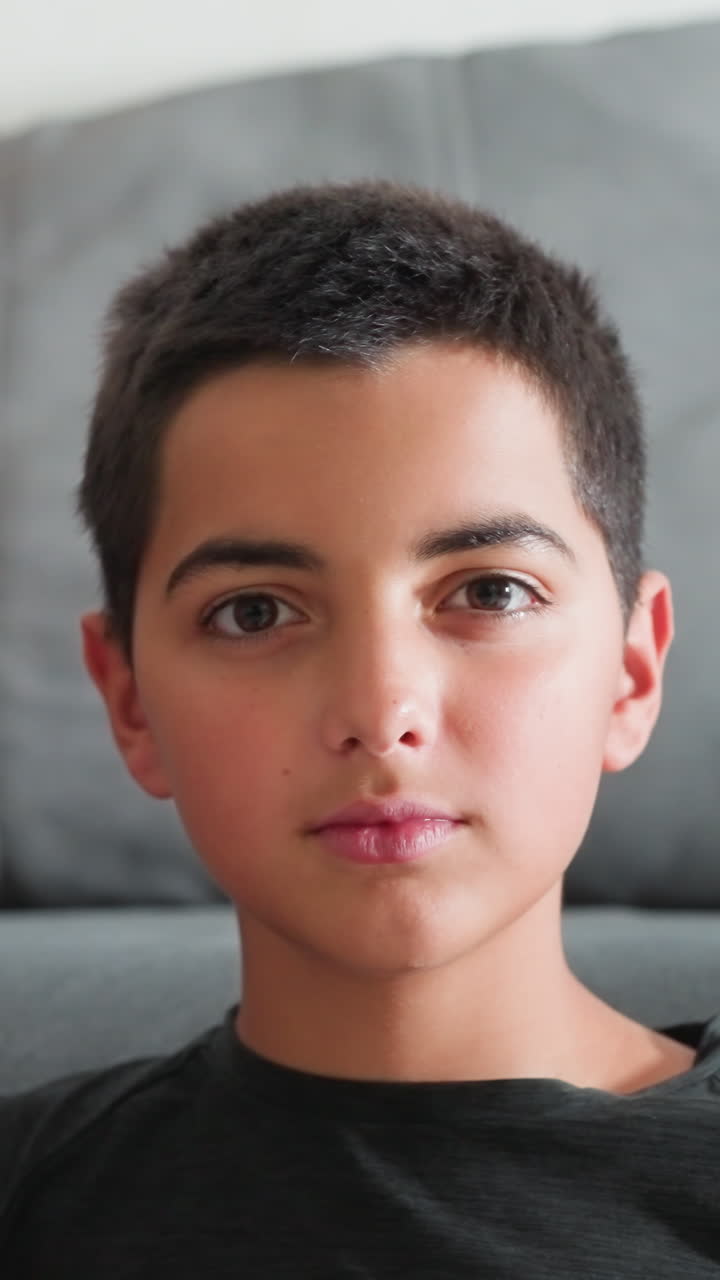 El hermano pequeño apoya la cabeza en el hombro de su hermana en tono divertido mientras ella sonríe con calidez; ambos miran directamente a la cámara sentados en el interior sobre un suave sofá gris, mostrando un dulce vínculo fraternal.