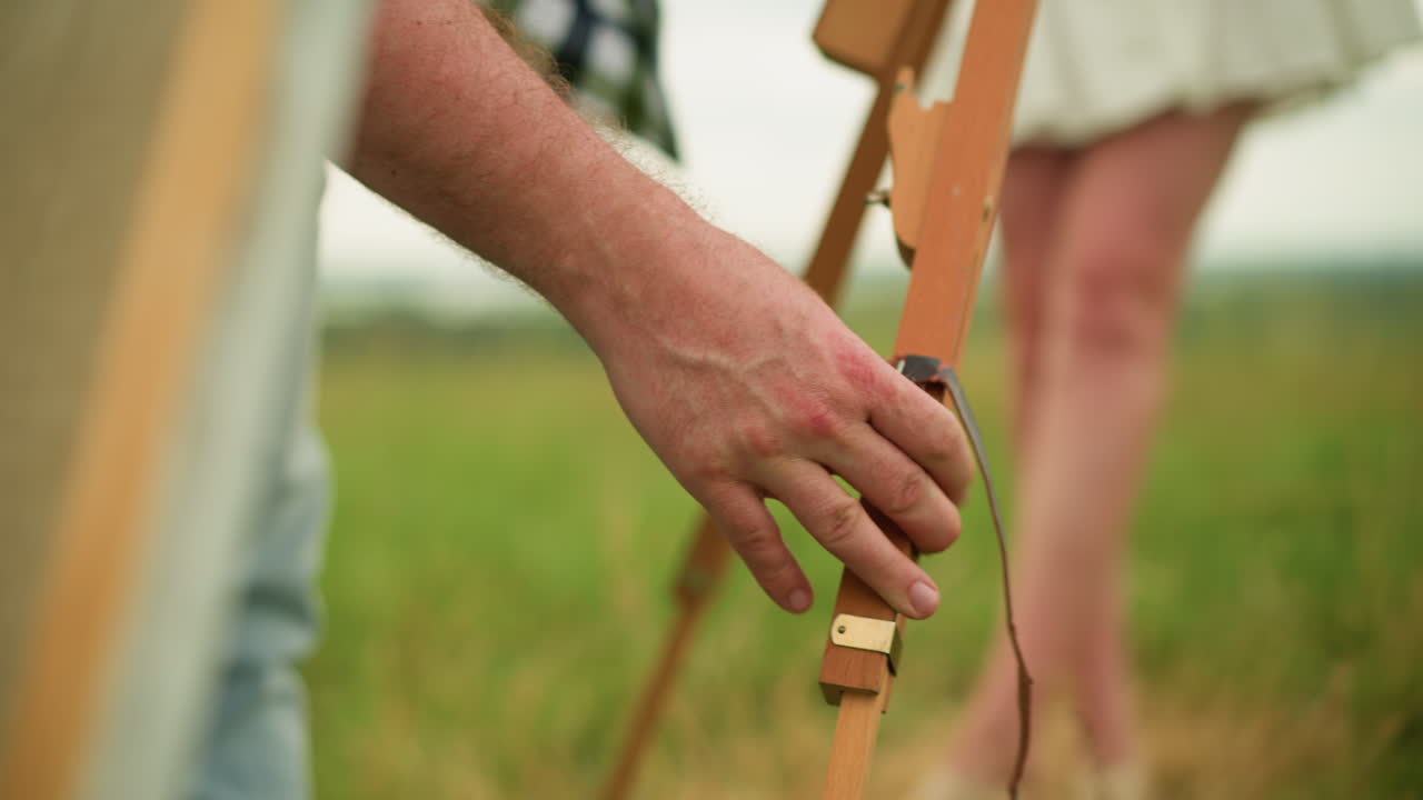 A close-up shot of a man's hand, dressed in a checked shirt, carefully adjusting a wooden tripod outdoors. A woman's leg is visible in the background