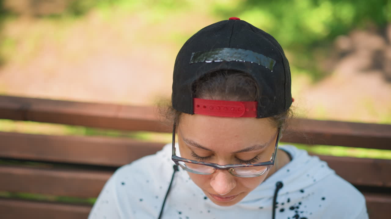 Joven sentado en un banco, reflexionando en silencio, con la mirada hacia arriba tras una pausa para tomar un tentempié, con gafas y gorra, con un fondo suave de follaje iluminado por el sol, con auriculares a la vista, sudadera informal, retrato contemplativo de tarde