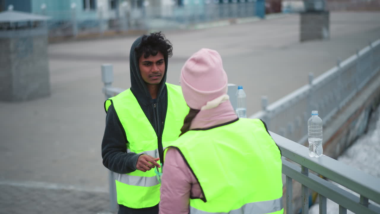 Volunteers in high visibility vests taking break on bridge during cold weather, standing near railing, talking and eating from food container, with water and ice visible in background, promoting teamwork