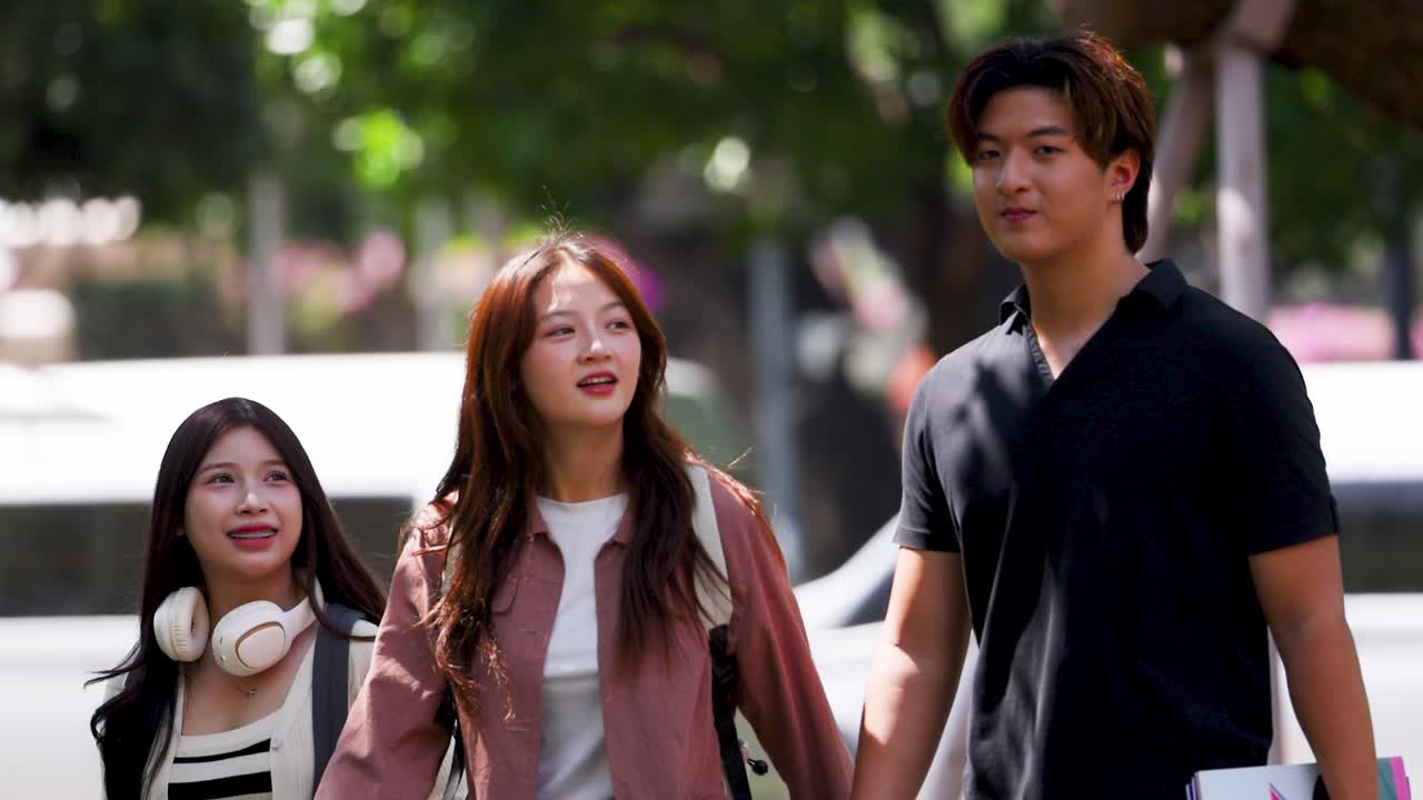 นักเรียนเอเชียรุ่นเยาว์สามคนเดินด้วยกันกลางแจ้งในวิทยาเขตที่มีแดดสดใสขณะพูดคุยและยิ้ม