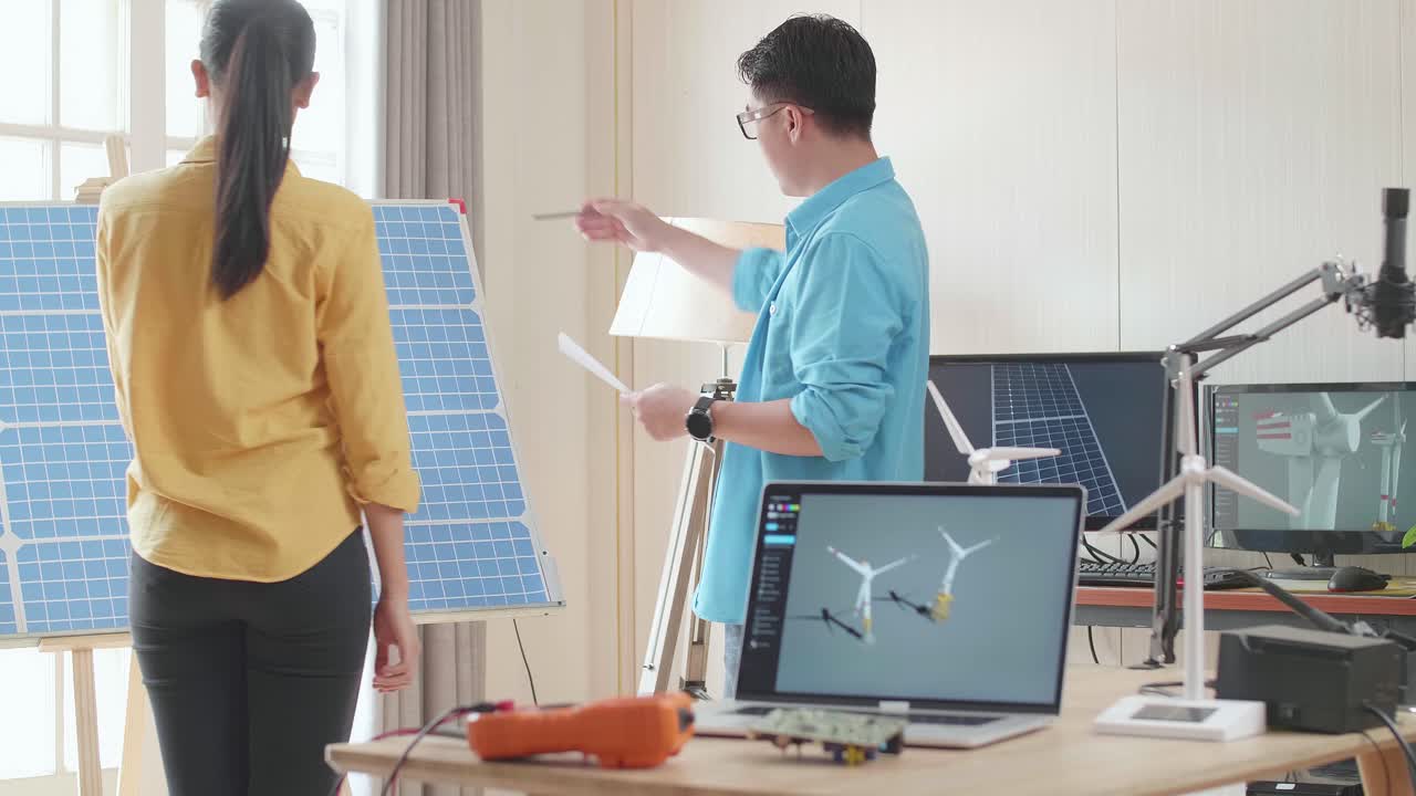Asian Man And Woman Discuss Work With Solar Cell Panel And Laptop Computer Showing Wind Turbine