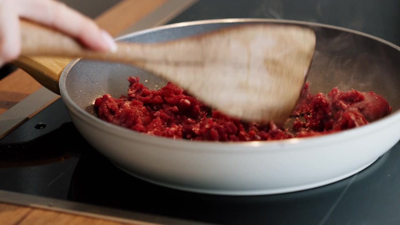 Close-up of breaking raw minced beef into smaller pieces with a wooden spatula in a frying pan, preparing the ingredient for cooking.