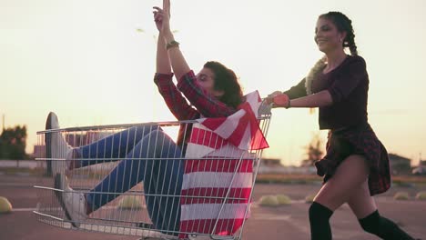 young hipster teen girls having fun at the shopping mall parking, riding in shopping cart holding the american flag