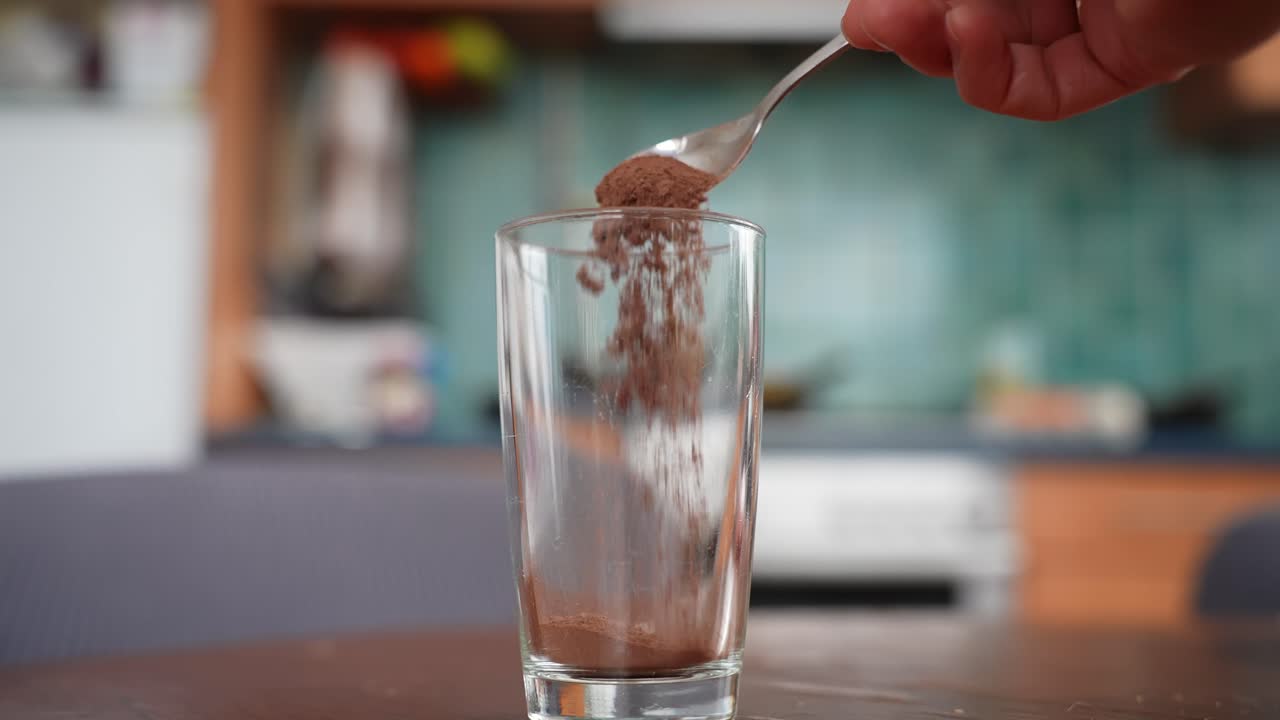 Una mano cosparge cacao in polvere da un cucchiaio in un bicchiere vuoto su un tavolo di legno, iniziando a preparare una cioccolata calda fatta in casa in un'accogliente cucina con un morbido effetto bokeh