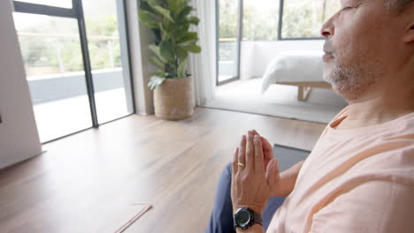 un anciano biracial practicando meditación de yoga en su casa.