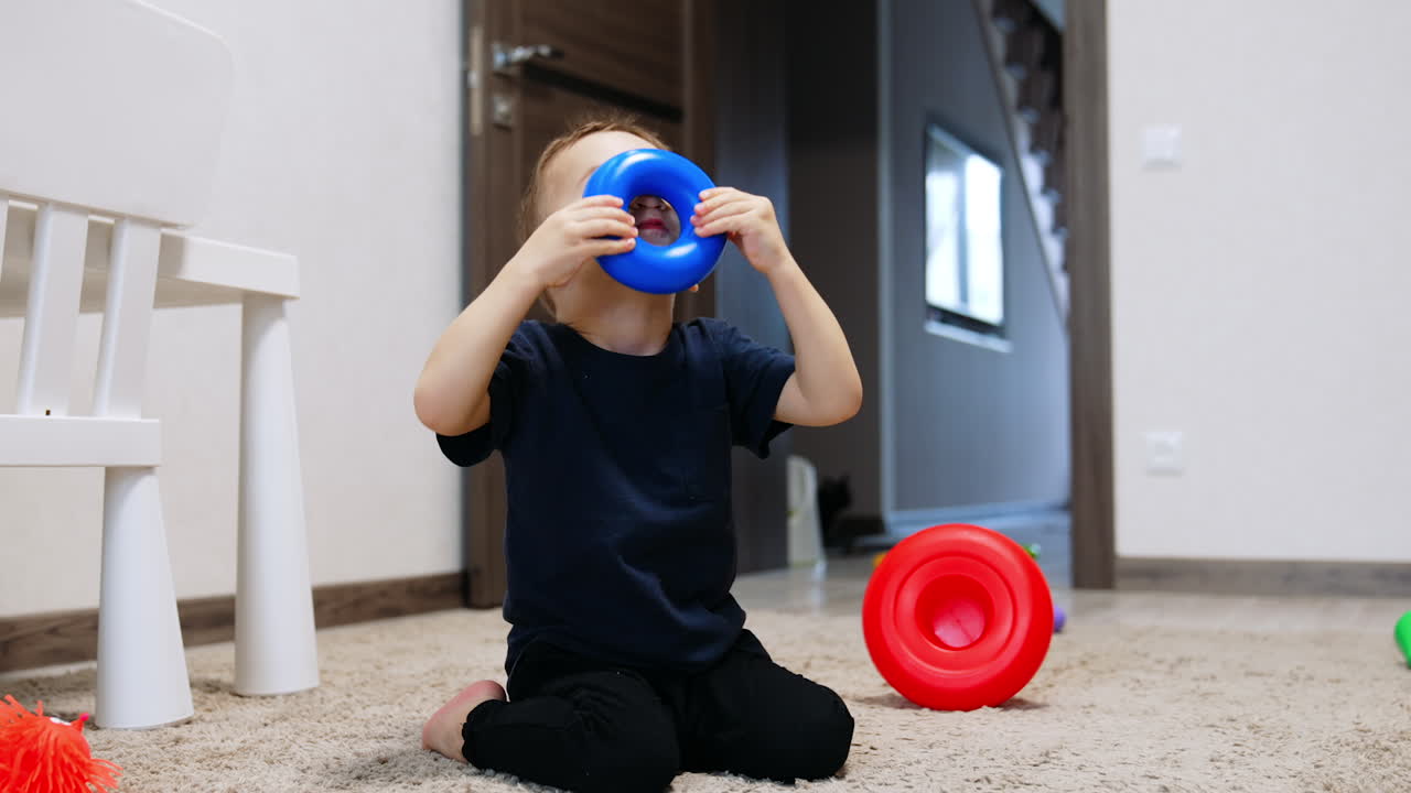 Baby boy wearing dark clothes sitting on the carpet. Kid holds the ring from pyramid at his face.