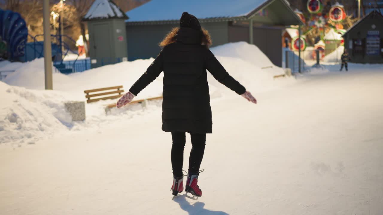Skater gliding smoothly on outdoor ice rink in winter evening, arms outstretched for balance, wearing black coat, red skates, patterned mittens, enjoying seasonal activity surrounded by snow